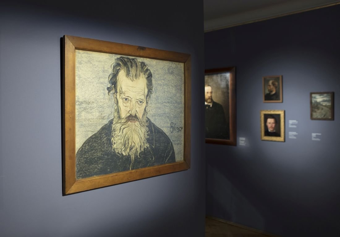 Szkicowany portret starszego mężczyzny z długą siwą brodą w drewnianej ramie na fioletowo-niebieskiej ścianie galerii, po prawej w tle widoczne mniejsze portrety i opisy ekspozycji.