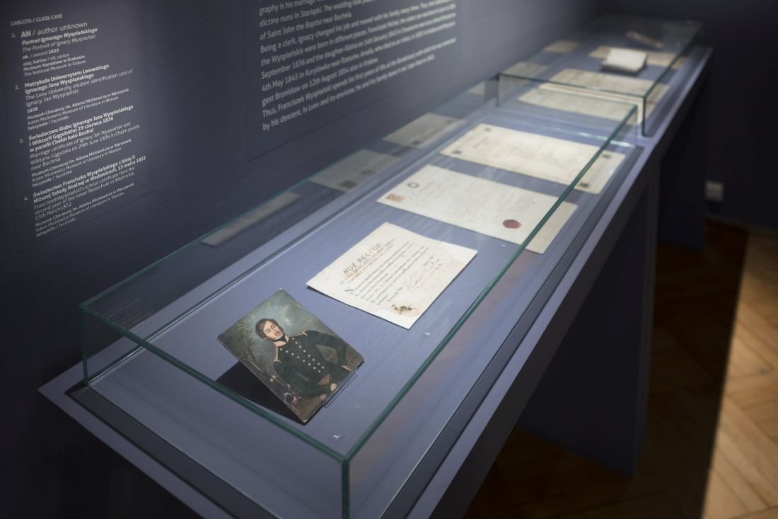 Przeszklona witryna muzealna ustawiona ukośnie, z niebieskim podkładem i kilkoma starymi dokumentami oraz małym portretem postaci w mundurze, z opisami na ciemnej ścianie i fragmentem drewnianej podłogi po prawej.