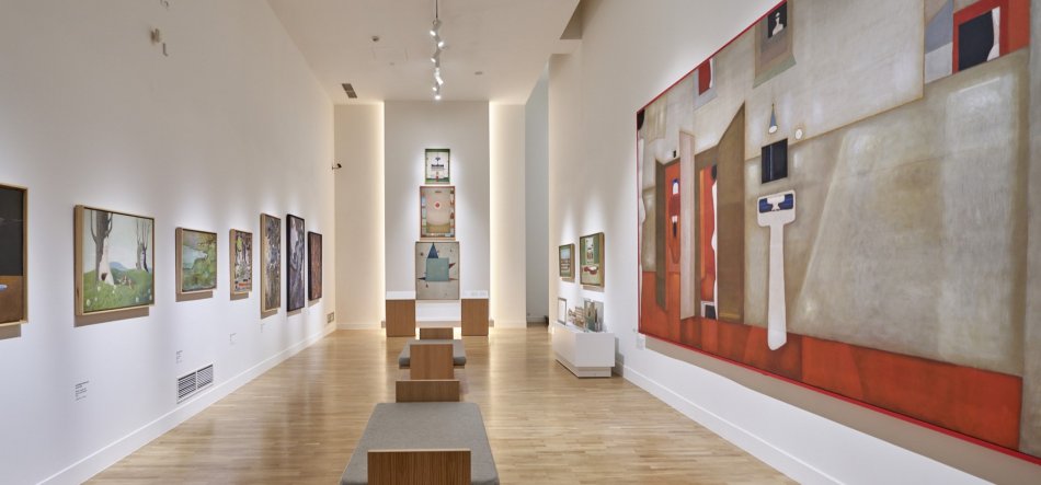 Wnętrze jasnej galerii z długą białą ścianą i drewnianą podłogą, widok w perspektywie na rząd mniejszych obrazów w ramach po lewej i duże czerwono‑beżowe dzieło po prawej, centralnie ustawione drewniane ławki z tapicerowanymi siedziskami oraz punktowe oświetlenie sufitowe, a na końcu podświetlona nisza z kilkoma zawieszonymi pracami.