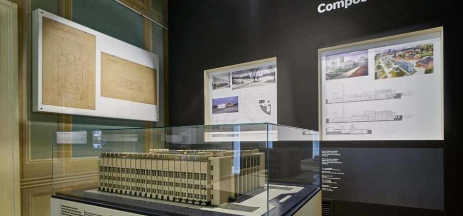Model architektoniczny prostokątnego budynku z rytmiczną elewacją umieszczony w szklanej gablocie na postumencie, wokół niego ściany wystawowe z oprawionymi rysunkami technicznymi, planami i wizualizacjami.