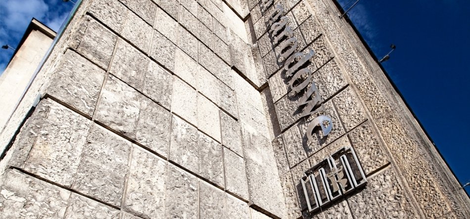 Zdjęcie z niskiego kąta przedstawiające narożnik dużego budynku wyłożony prostokątnymi, szorstkimi betonowymi panelami z pionowo przymocowanymi metalowymi literami na elewacji, po prawej jasne niebo z kilkoma chmurami.