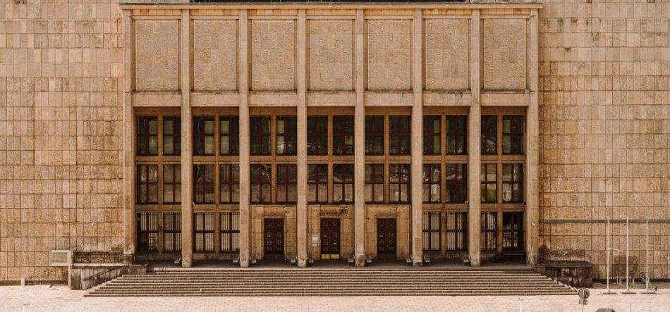 Symetryczna elewacja dużego beżowego budynku o geometrycznym układzie: wysokie pionowe pilastry dzielą rząd dużych prostokątnych okien z licznymi podziałami nad trzema ciemnymi wejściami, przed którymi ciągną się szerokie schody i otwarty, wyłożony płytami plac.