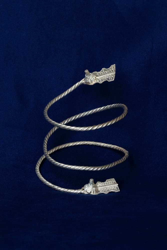 Srebrzysty, skręcony metalowy drut uformowany w pionową spiralę z trzema zwojami i ozdobnymi reliefowymi zakończeniami na obu końcach, ustawiony na gładkim ciemnoniebieskim tle.