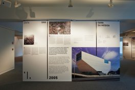 Duży panel wystawowy z białych tablic informacyjnych i zdjęć ustawiony w pomieszczeniu wystawowym, z sufitowymi reflektorami i szarą dywanową podłogą; po prawej duże zdjęcie nowoczesnego białego, kanciastego budynku na tle błękitnego nieba, po lewej mniejsze fotografie (w tym zdjęcie z wieloma osobami) oraz bloki tekstu zawierające widoczne daty „1999” i „2005”.