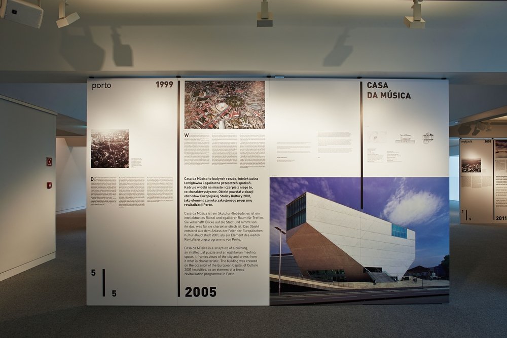 Trzy panele wystawowe w jasnym wnętrzu muzealnym z szarym dywanem i sufitowymi reflektorami: lewy panel z napisem "porto 1999" i małym zdjęciem, środkowy z blokiem tekstu, zdjęciem z lotu ptaka i datą "2005", prawy z dużym zdjęciem nowoczesnego białego budynku i tytułem "CASA DA MÚSICA".