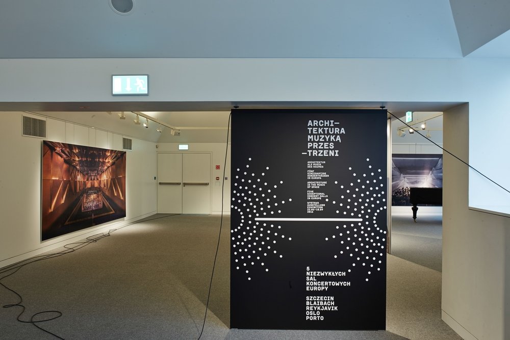 Wnętrze galerii z centralną czarną ścianką z białymi kropkami i tekstem po polsku „ARCHITEKTURA MUZYKA PRZESTRZENI”, po lewej duża fotografia sali koncertowej i leżące kable na szarym dywanie, w głębi podwójne drzwi z zielonym znakiem ewakuacyjnym, a po prawej fragment fortepianu i kolejna fotografia na ścianie.
