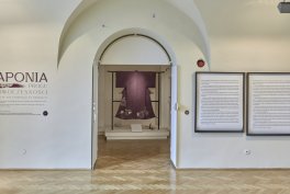 W jasnej galerii z drewnianą podłogą łukowate przejście prowadzi do wnęki z wystawioną fioletową szatą na stojaku; po lewej fragment ściennego napisu, po prawej dwie oprawione plansze tekstowe i czerwony przycisk alarmowy.