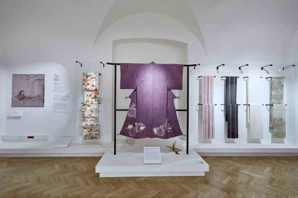 Galeria muzealna z białymi ścianami i łukowym sufitem, na centralnym czarnym stojaku wystawione fioletowe kimono z jasnymi, kwiatowymi plamami, po lewej w szklanych gablotach kolorowe ozdobne pasy i plansza z ilustracją i opisem, po prawej w oszklonych wnękach zawieszone pionowe tekstylne panele, całość na białej podstawie nad drewnianym parkietem.