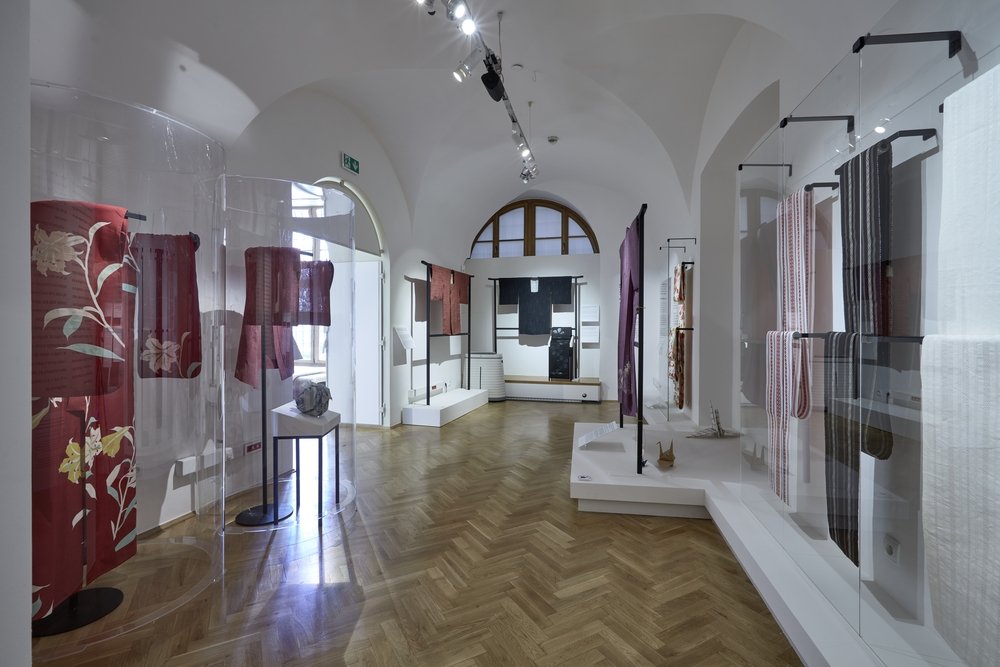 Wnętrze białej galerii z łukowatym sufitem i drewnianą podłogą w jodełkę, z ekspozycją zawieszonych i ułożonych tkanin i szat prezentowanych na stojakach oraz w przezroczystych cylindrycznych gablotach po lewej i w przeszklonych witrynach po prawej, oświetlone reflektorami i z dużym półokrągłym oknem w tylnej ścianie.