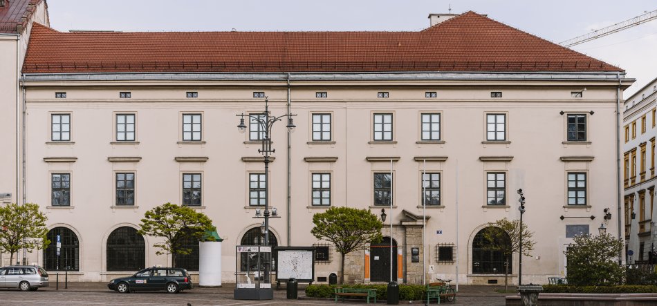 Frontowy widok na symetryczny, trzykondygnacyjny jasnobeżowy budynek z czerwoną dachówką, prostokątnymi oknami na dwóch górnych kondygnacjach i dużymi łukowymi oknami na parterze, a przed nim chodnik z kilkoma niskimi drzewami, ławkami, latarnią uliczną, przeszklonym kioskiem z mapą oraz zaparkowanymi samochodami.