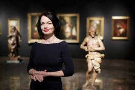 Kobieta o krótkich ciemnych włosach i czerwonych ustach, w czarnej sukience z rękawami do łokcia, stoi złożonymi dłońmi i delikatnie się uśmiecha na tle ciemnej sali wystawowej z dwiema rzeźbami na podestach i trzema oprawionymi obrazami na ścianie.