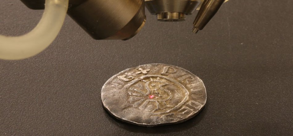Zużyta metalowa moneta leżąca na ciemnej matowej powierzchni, nad nią części aparatury optycznej (obiektywy i przewód), a na środku monety widoczna mała czerwona kropka lasera.