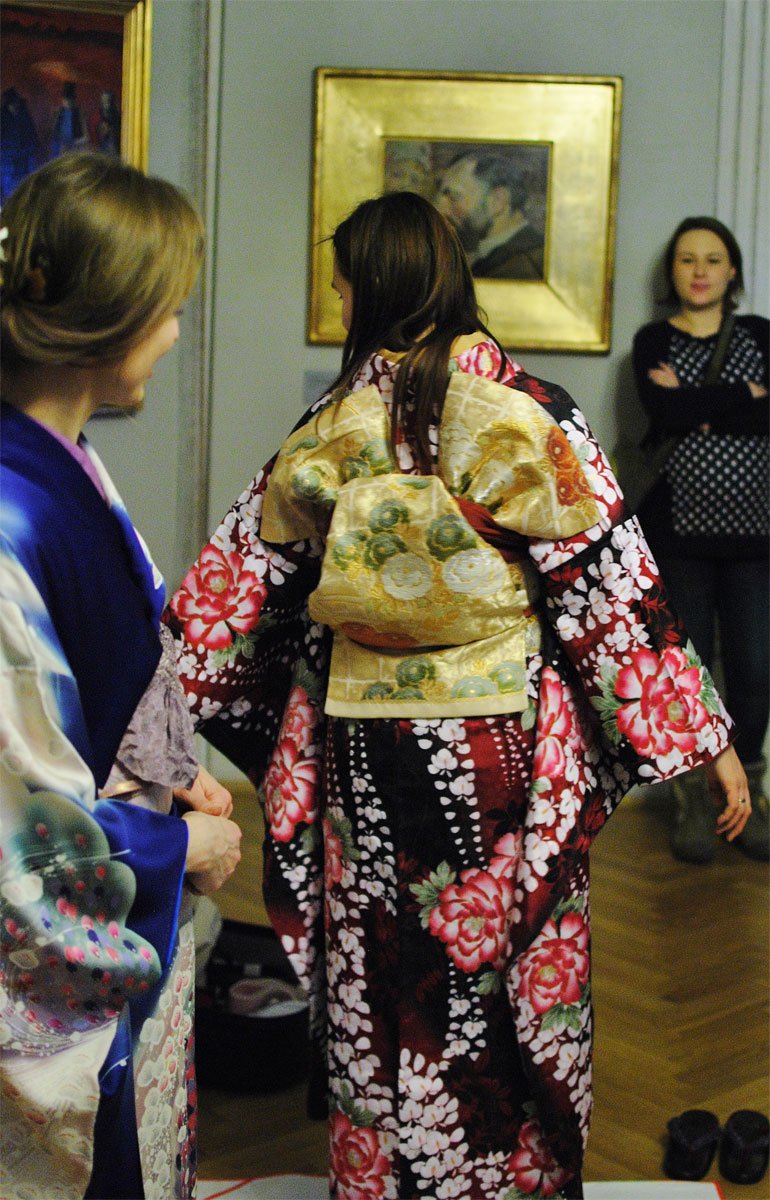 W środku kadru kobieta widoczna od tyłu w kolorowym kimono w duże czerwone kwiaty z szerokim złotym pasem zawiązanym z tyłu, po lewej częściowo widoczna inna osoba w niebieskim kimono, w tle na ścianie obraz w złotej ramie i stojąca z założonymi rękami kobieta w zachodnim ubraniu, na drewnianej podłodze para butów.