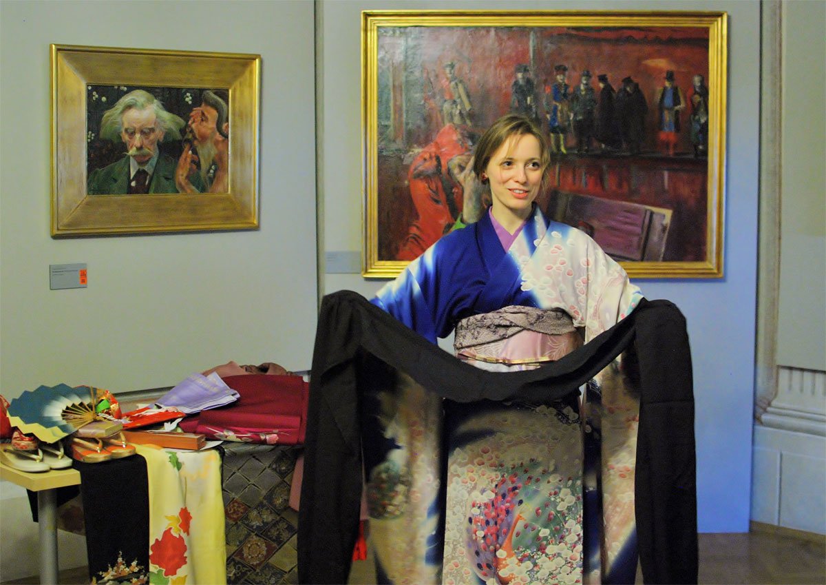 Kobieta w kolorowym kimonie stoi w sali wystawowej przed dwoma obrazami w złotych ramach, rozkładając czarny pas materiału, obok stołu z wachlarzami, złożonymi tkaninami i ozdobnymi klapkami.