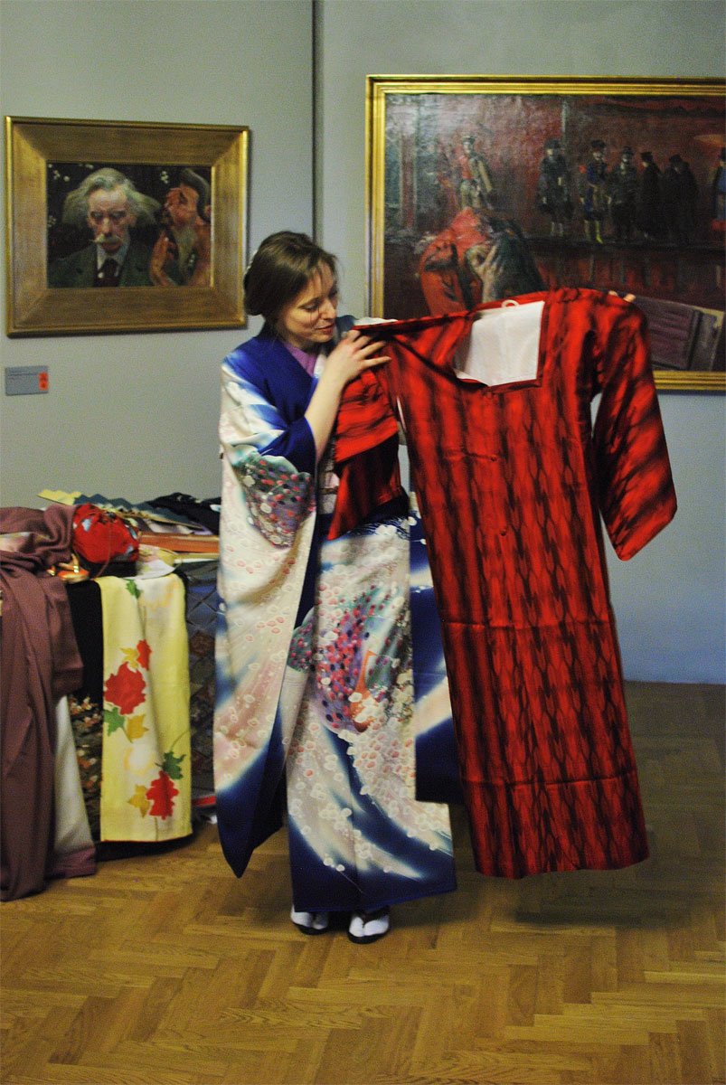 Kobieta ubrana w ozdobne niebiesko‑białe kimono stoi na drewnianej podłodze przy ścianie z oprawionymi obrazami i trzyma na wieszaku czerwone kimono, obok znajduje się stół z innymi tkaninami.