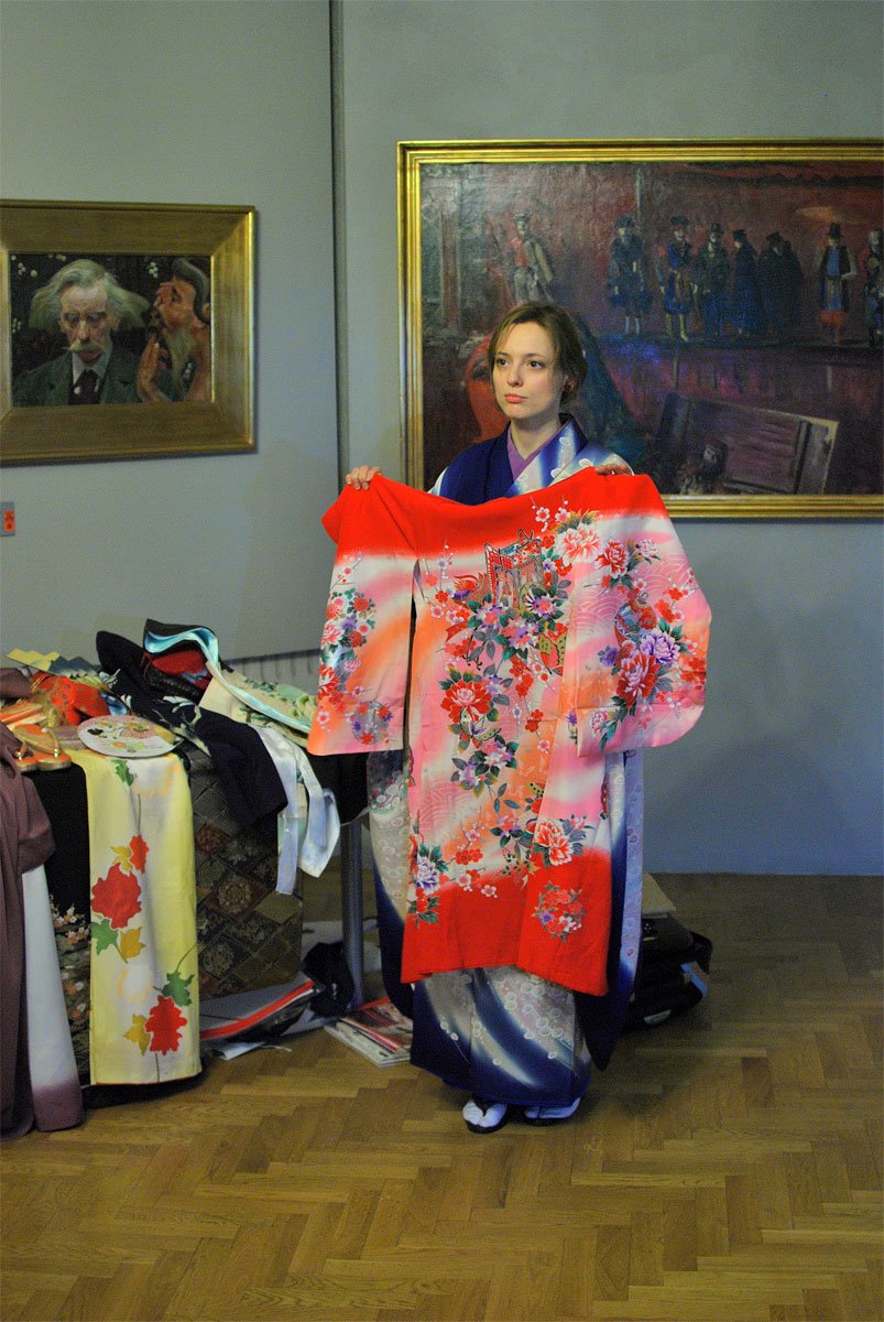 Kobieta stojąca na drewnianym parkiecie w sali wystawowej trzyma rozprostowane czerwono‑różowe kimono z bogatym kwiatowym wzorem; po lewej widoczny stół z innymi tkaninami i akcesoriami, a za nią na ścianie wiszą dwa duże obrazy w złotych ramach.