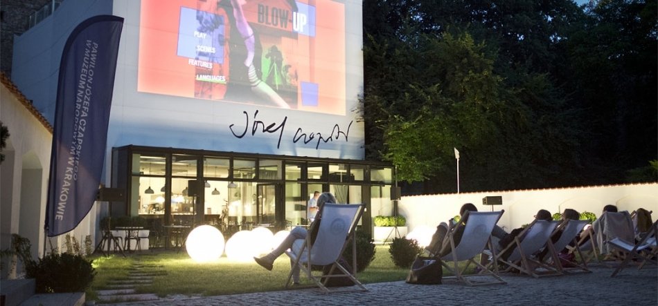 Wieczorny plenerowy pokaz filmowy przed nowoczesnym budynkiem z przeszklonym parterem, dużym prostokątnym ekranem projekcyjnym na białej ścianie, rzędem leżaków z widzami po prawej, kilkoma podświetlonymi kulami na trawie i wysoką flagą reklamową po lewej, w tle ciemne drzewa.
