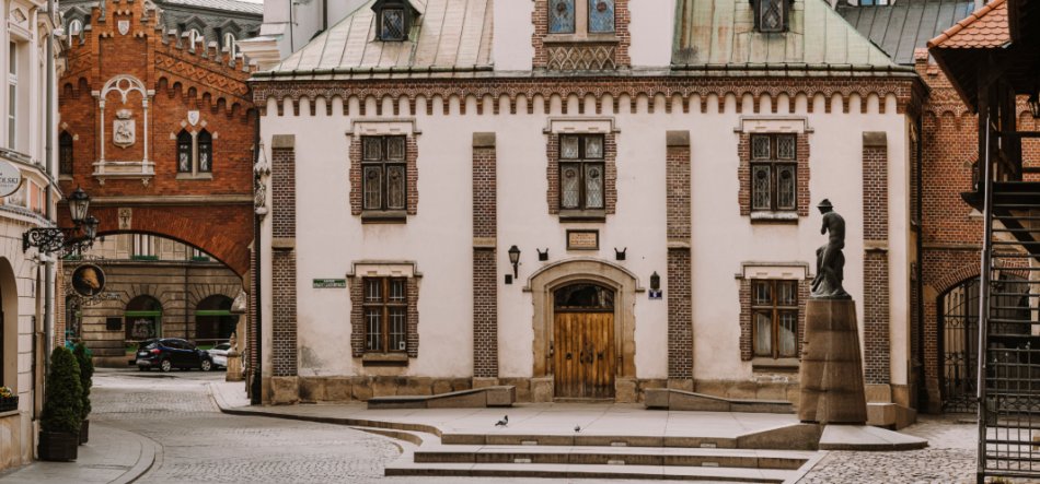 Brukowana ulica i niewielki plac z kilkoma stopniami przed białą kamienicą z ceglaną oprawą okien, drewnianymi drzwiami i zielonkawym dachem, obok której stoi figura na wysokim cokole; z lewej czerwony ceglasty łuk prowadzący do podwórza, w tle samochód, latarnie i kilka gołębi.