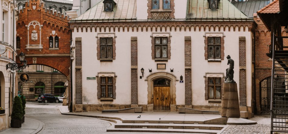 Plac z brukowanym chodnikiem i niewielkimi schodami przed zabytkowym budynkiem o białej elewacji z ceglaną dekoracją, centralnymi drewnianymi drzwiami i ozdobnymi oknami; po prawej rzeźba na wysokim cokole, po lewej łuk ceglanej bramy prowadzący do bocznej uliczki, na placu gołębie, latarnie i fragmenty sąsiednich kamienic.