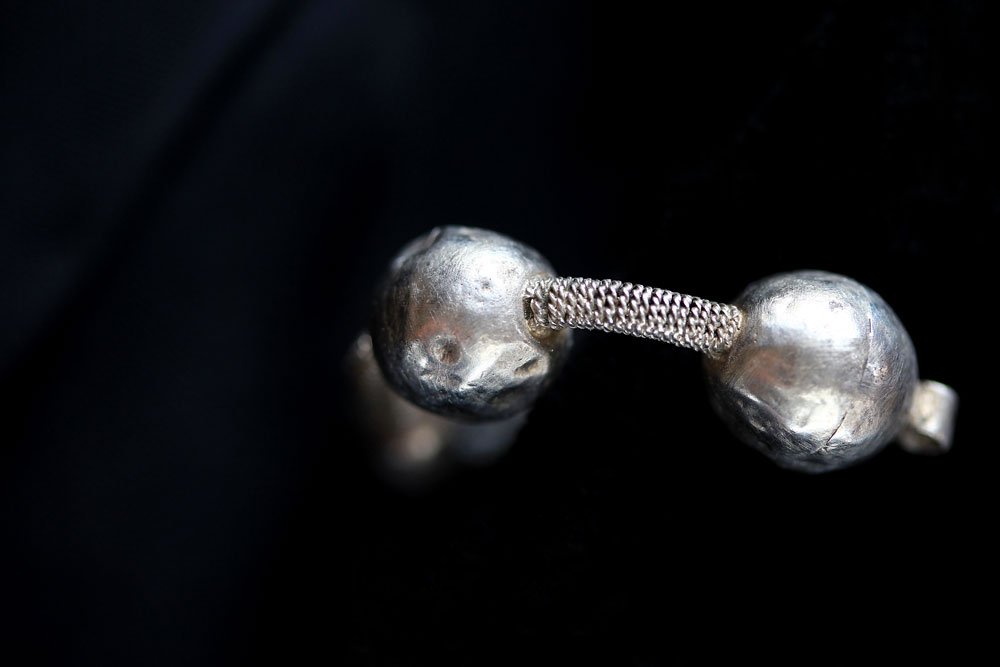 Zbliżenie na srebrzysty metalowy przedmiot składający się z dwóch kul o młotkowanej powierzchni połączonych krótkim, plecionym łącznikiem, na gładkim czarnym tle.