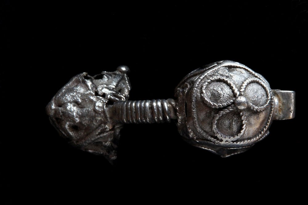 Srebrny metalowy przedmiot na czarnym tle z dwoma ozdobnymi końcami połączonymi żebrowanym trzpieniem: po prawej kulisty element z trzema wypukłymi okręgami ozdobionymi ziarnistym konturem i małym guzkiem, po lewej nieregularna pofałdowana kula z otworami.