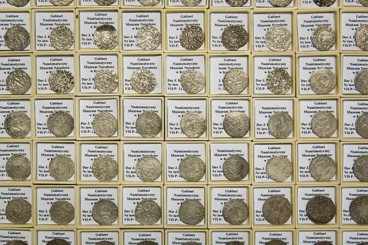 Widok z góry na równą siatkę kremowych pudełek z białymi etykietami z czarnym drukiem (w tym widocznym napisem „Gabinet Numizmatyczny Muzeum Narodowe”) i pojedynczą srebrzystą monetą umieszczoną na każdym pudełku.