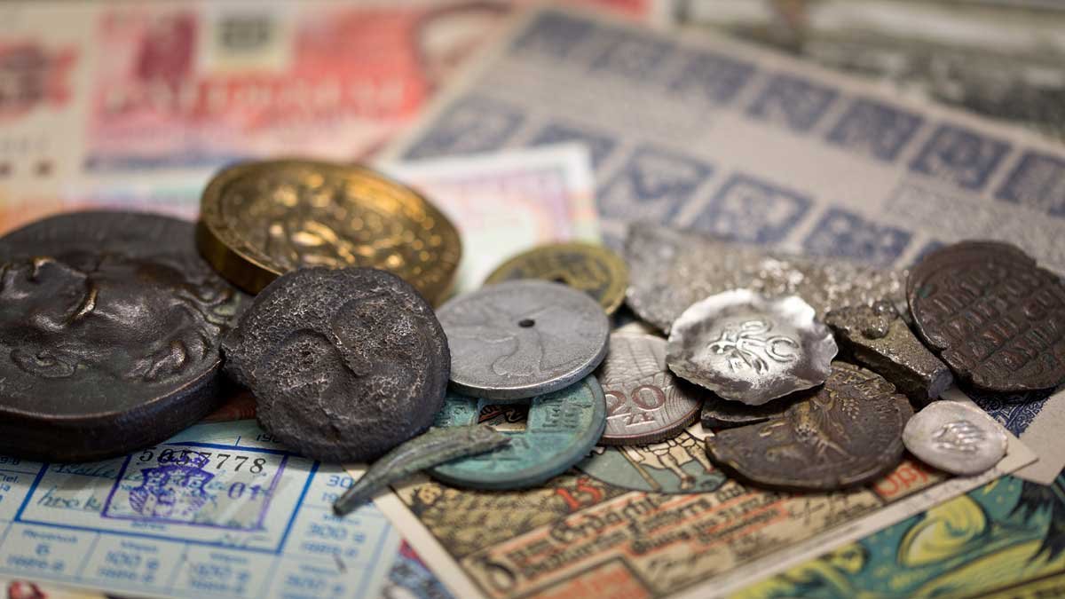 Zbliżenie na rozrzucone na banknotach różnokolorowe, zużyte monety i krążki o różnych kształtach, rozmiarach i fakturach, z widoczną patyną, wybitymi wzorami i niewielkimi otworami, na płytko rozmytym tle.