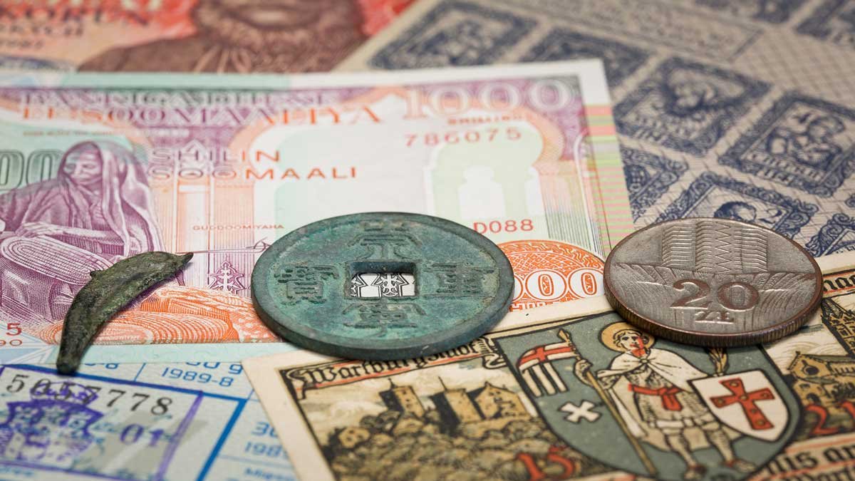 Różne banknoty i monety rozłożone na płaskiej powierzchni — na środku zielonkawa okrągła moneta z kwadratowym otworem, obok metalowa moneta z nominałem 20, mały zakrzywiony metalowy kawałek oraz kolorowy banknot 1000 soomaali i karta z herbem.