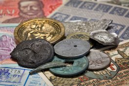 Kilka metalowych monet różnych rozmiarów i kolorów (ciemne, zielonkawe, srebrne i złote) ułożonych na banknotach i papierowych dokumentach z fragmentami nadruków i pieczątkami.