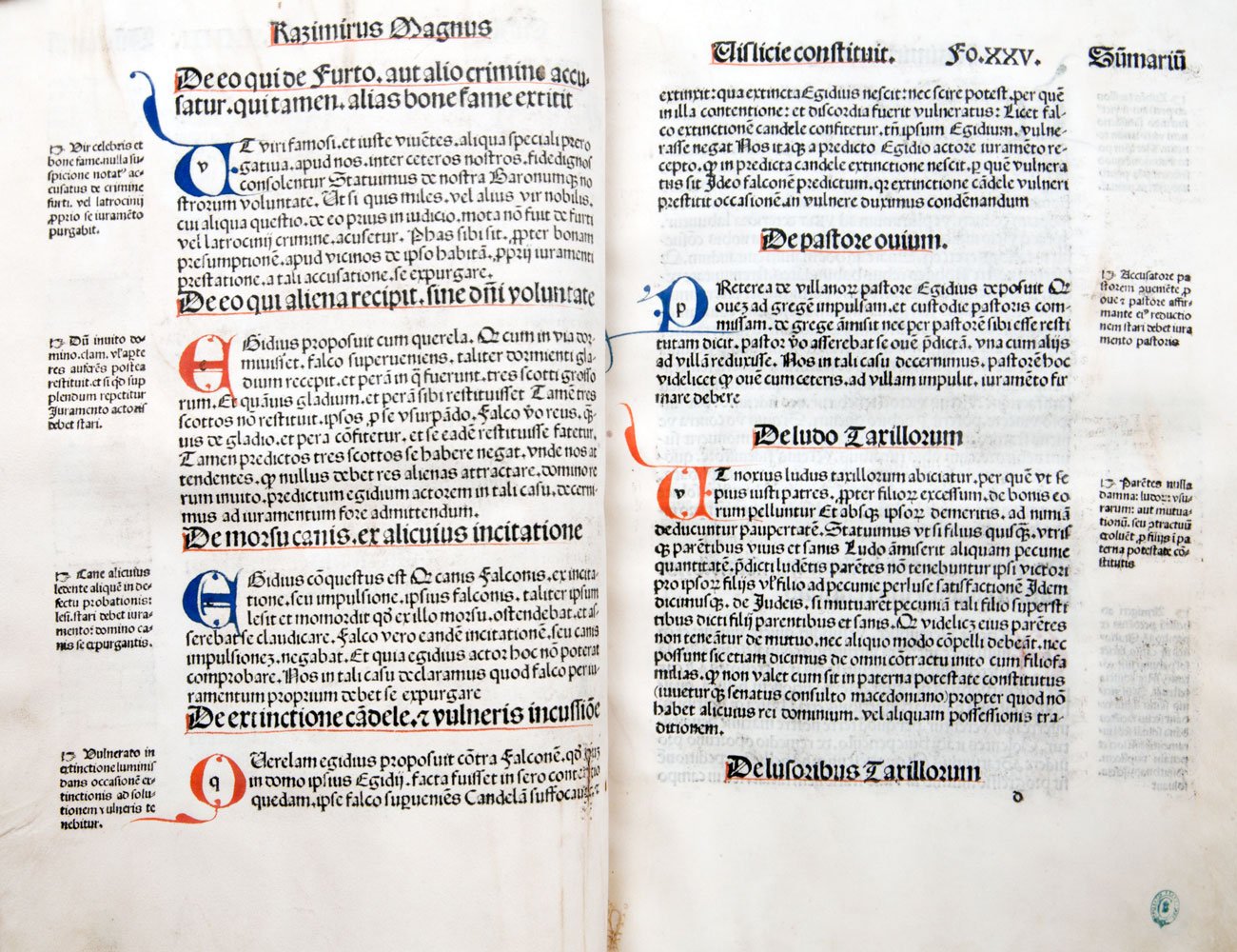 Dwustronicowy rozkład starego druku z gęstym czarnym tekstem w stylu gotyckim, kolorowymi czerwonymi i niebieskimi inicjałami, nagłówkami, wąskimi kolumnami i marginaliami, śladem zszycia w środku oraz małą zieloną pieczęcią w prawym dolnym rogu.