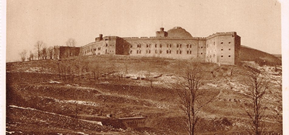 Sepiowana fotografia panoramiczna przedstawiająca masywny kamienny budynek warowny o długim, trójkondygnacyjnym korpusie z regularnymi oknami i zaokrąglonymi basztami na szczycie łagodnego, częściowo zaśnieżonego wzgórza, przed którym rozciągają się tarasowane pola i kilka nagich drzew.