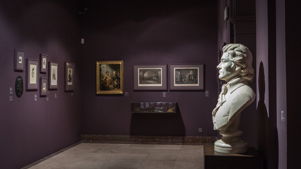 Wnętrze galerii z ciemnofioletowymi ścianami, po prawej na pierwszym planie białe popiersie mężczyzny na cokole rzucające cień na ścianę, w tle po lewej i na środkowej ścianie oprawione rysunki i obrazy oraz przeszklona witryna ekspozycyjna na niskim cokole.
