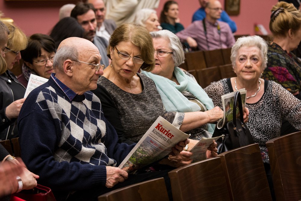 Grupa starszych osób siedzi w rzędach drewnianych krzeseł; na pierwszym planie starszy mężczyzna w okularach i granatowym swetrze w romby trzyma gazetę „Kurier”, obok dwie kobiety z okularami — jedna rozmawia i patrzy na gazetę, druga trzyma torebkę i broszurę, a w tle widać więcej siedzących osób.
