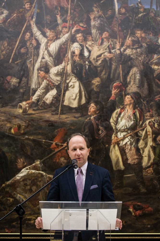 Mężczyzna w ciemnym garniturze i różowym krawacie stoi przy mikrofonie za przezroczystym pulpitem na tle dużego historycznego obrazu przedstawiającego licznych postaci z bronią i uniesionymi rękami.