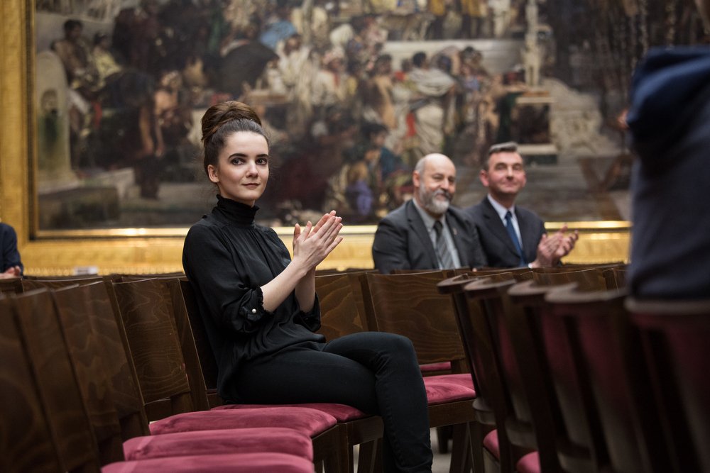 Młoda kobieta w czarnym ubraniu i włosach upiętych w kok siedzi i klaszcze na drewnianym krześle z czerwonym siedziskiem, w tle duży obraz w złotej ramie oraz dwaj mężczyźni w garniturach także klaszczący, a po prawej widać rzędy pustych drewnianych krzeseł.