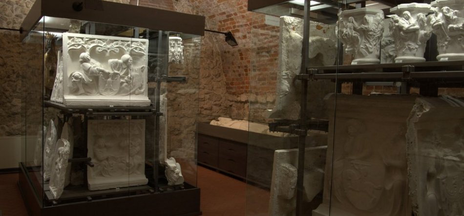 Wnętrze wystawowe z ceglanymi ścianami i sklepieniem, w którym szklane gabloty z metalowymi stelażami prezentują białe gipsowe odlewy reliefów i fragmenty rzeźb oraz niska witryna z płaskimi kamiennymi płytami, o ciepłym, przytłumionym oświetleniu.