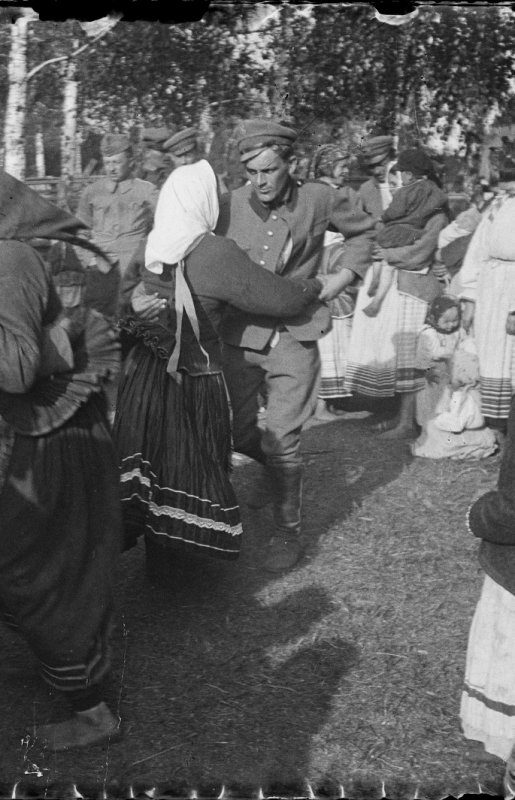 Czarno-białe zdjęcie plenerowe grupy ludzi tańczących; centralnie mężczyzna w mundurze trzymający kobietę w białej chustce i ciemnej, zdobionej spódnicy, wokół zgromadzenie osób w tradycyjnych strojach i dzieci, w tle drzewa i trawa.