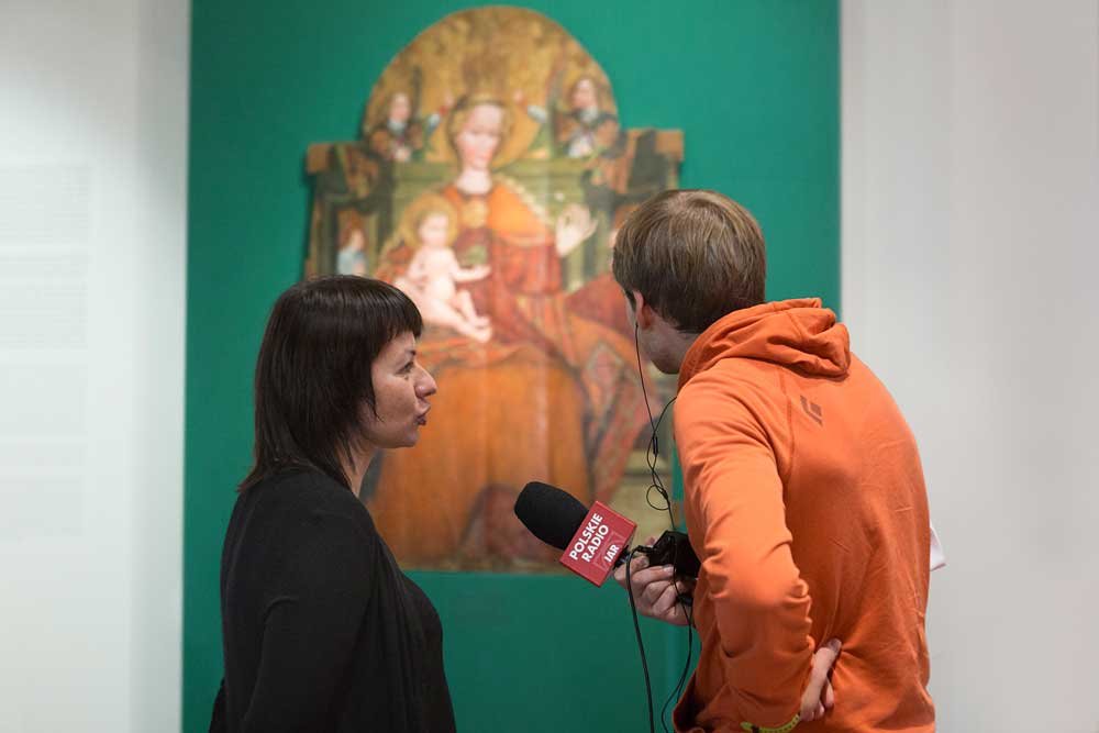 Kobieta z krótkimi ciemnymi włosami w czarnym ubraniu stoi bokiem i rozmawia z mężczyzną w pomarańczowej bluzie, który trzyma mikrofon z napisem Polskie Radio i urządzenie nagrywające; w tle, w galerii, widoczny zielony panel z obrazem przedstawiającym kobietę z dzieckiem.