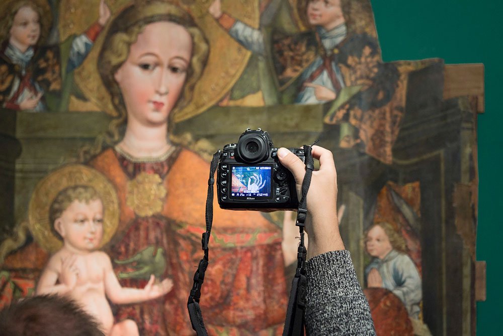Zbliżenie osoby w szarym swetrze trzymającej lustrzankę skierowaną na duży obraz sakralny przedstawiający kobietę z aureolą i nagie dziecko, na tylnym wyświetlaczu aparatu widać podgląd fotografowanej sceny.