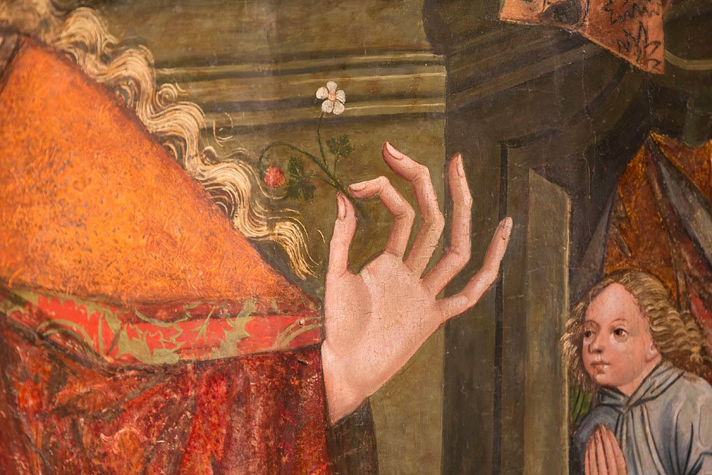 Fragment obrazu pokazujący zbliżenie dłoni z wyprostowanymi palcami trzymającej mały biały kwiatek; po lewej widoczny bogato zdobiony czerwono‑pomarańczowy rękaw i pasma jasnych falistych włosów, po prawej mała postać z lokami i złożonymi rękami.