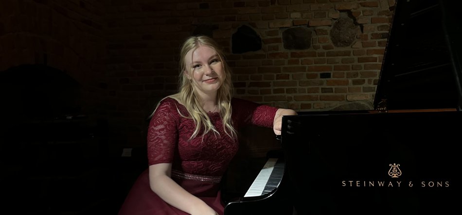 Kobieta o długich blond włosach w bordowej koronkowej sukni siedzi przy czarnym fortepianie Steinway & Sons na tle ceglanej ściany, słabo oświetlona, z ręką opartą na rancie instrumentu i delikatnym uśmiechem.