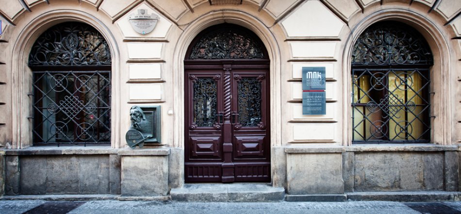Fasada kamienicy z trzema łukowymi otworami: pośrodku ciemnobrązowe, ozdobne dwuskrzydłowe drzwi z rzeźbionymi panelami i ażurowym nadświetlem, po obu stronach okna z żeliwnymi, wzorzystymi kratami, jasnobeżowa oprawa kamienna, po lewej metalowa płaskorzeźba z medalionem, po prawej tablica informacyjna, kamienna podmurówka i brukowany chodnik.