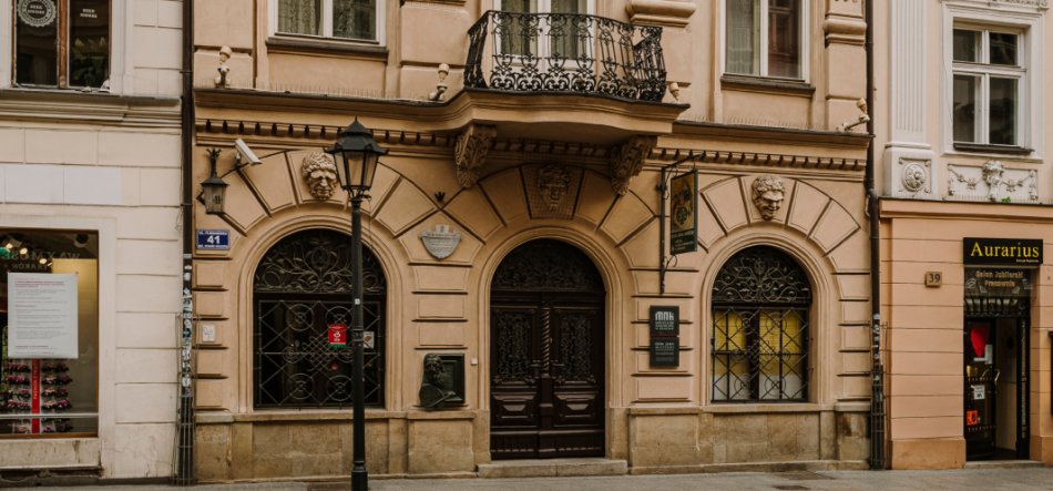 Fasada ozdobnego beżowo‑różowego budynku z centralnymi ciemnymi drewnianymi drzwiami w łukowym portalu, dwiema arkadowymi oknami z kutą kratą, żeliwnym balkonem nad wejściem, rzeźbionymi maskami nad arkadami, stojącą latarnią uliczną na chodniku i sklepami po bokach z widocznymi numerami 41 i 39.