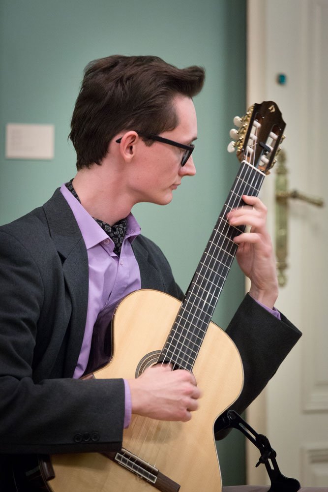 Mężczyzna w okularach i szarej marynarce, w liliowej koszuli, siedzi i gra na gitarze klasycznej, lewą ręką przy gryfie, prawą nad pudłem rezonansowym, na tle jasnozielonej ściany i białych drzwi z mosiężnym uchwytem.
