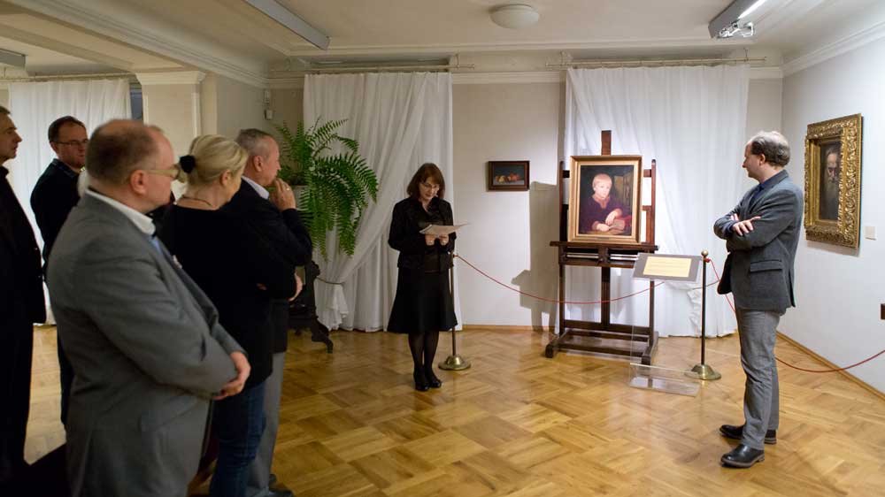 Wnętrze galerii z grupą zwróconych do siebie osób po lewej, kobietą czytającą z kartki przy sztaludze z oprawionym portretem, mężczyzną stojącym z założonymi rękami po prawej, czerwoną linką ochronną, parkietem, białymi zasłonami i dodatkowymi obrazami na ścianach.