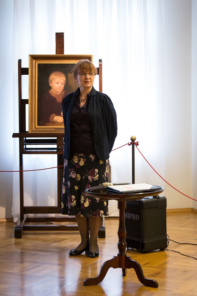 Kobieta w okularach w ciemnej marynarce i spódnicy w kwiaty stoi na drewnianej podłodze przed sztalugami z ramowym portretem dziecka, obok niski okrągły stolik z notatkami i mikrofonem oraz czarny głośnik oznaczony „MIPRO”, w tle białe zasłony i czerwony sznurek ochronny na słupkach.