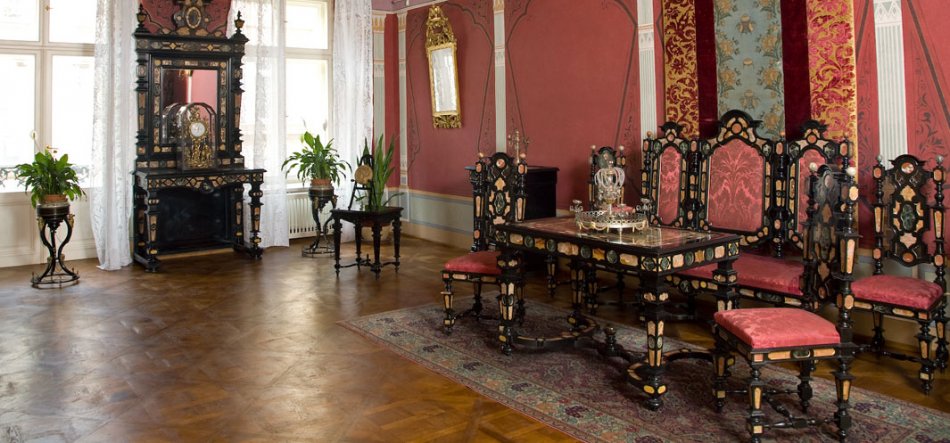 Widok eleganckiego, historycznego wnętrza z parkietową podłogą: po prawej czarny, bogato zdobiony stół otoczony wysokimi krzesłami z czerwonymi obiciami ustawionymi na wzorzystym dywanie, za nimi dekoracyjne czerwono‑złote pasy tkaniny na ścianie; po lewej duże okna z białymi koronkowymi zasłonami oraz wysoka czarna, zdobiona szafa z lustrem i zegarem, a w pomieszczeniu także rośliny doniczkowe na metalowych stojakach i złocone wiszące lustro.