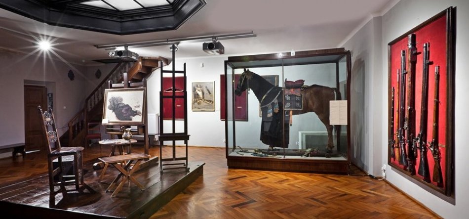 Panoramiczne wnętrze sali muzealnej z parkietową podłogą: po lewej drewniane podium z ozdobnym rzeźbionym krzesłem, małym stolikiem i sztalugą z oprawionym obrazem głowy zwierzęcia oraz schodami w tle; centralnie i po prawej duża szklana witryna z wypchanym koniem w uprzęży i siodle z tabliczką informacyjną; na prawej ścianie ekspozycja kilku długich broni palnych na czerwonym tle, sufitowe reflektory oświetlają salę.