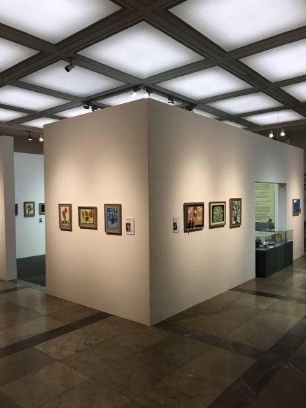 Wnętrze galerii z białą narożną ścianką w środku, na której w rzędzie wiszą małe kolorowe obrazy w ramach, oświetlone punktowymi lampami pod kwadratowym, podświetlanym sufitem, na polerowanej kamiennej posadzce, z gablotami i przejściami w tle.