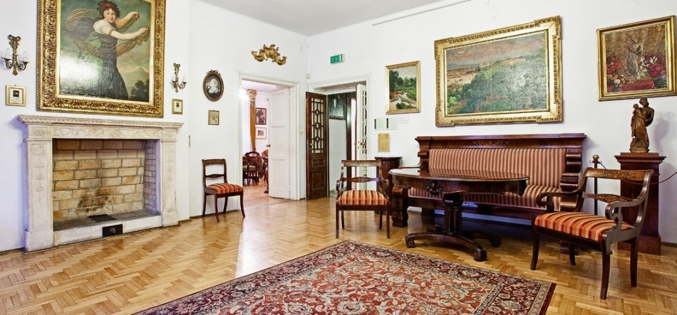 Wnętrze eleganckiego pokoju z jasnym drewnianym parkietem i wzorzystym dywanem na pierwszym planie; po lewej kamienny kominek z dużym, złoconym obrazem nad nim, na ścianach kilka oprawionych płócien i kinkietów, w centrum otwarte drzwi z zielonym znakiem wyjścia nad nimi, a po prawej antyczna drewniana sofa i krzesła z czerwono‑pasiastym obiciem oraz rzeźba na postumencie.